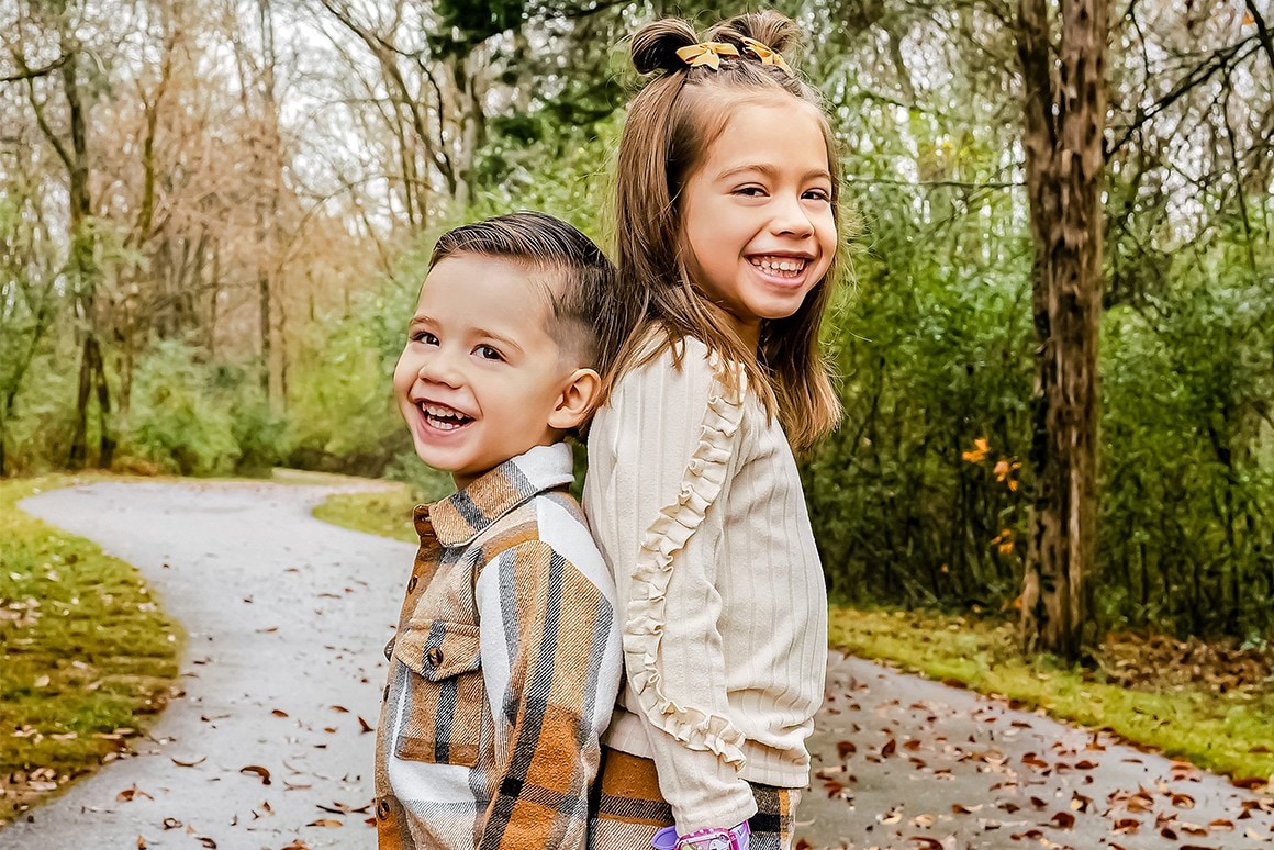 Brother and sister laughing back-to-back on a leaf-covered path in fall